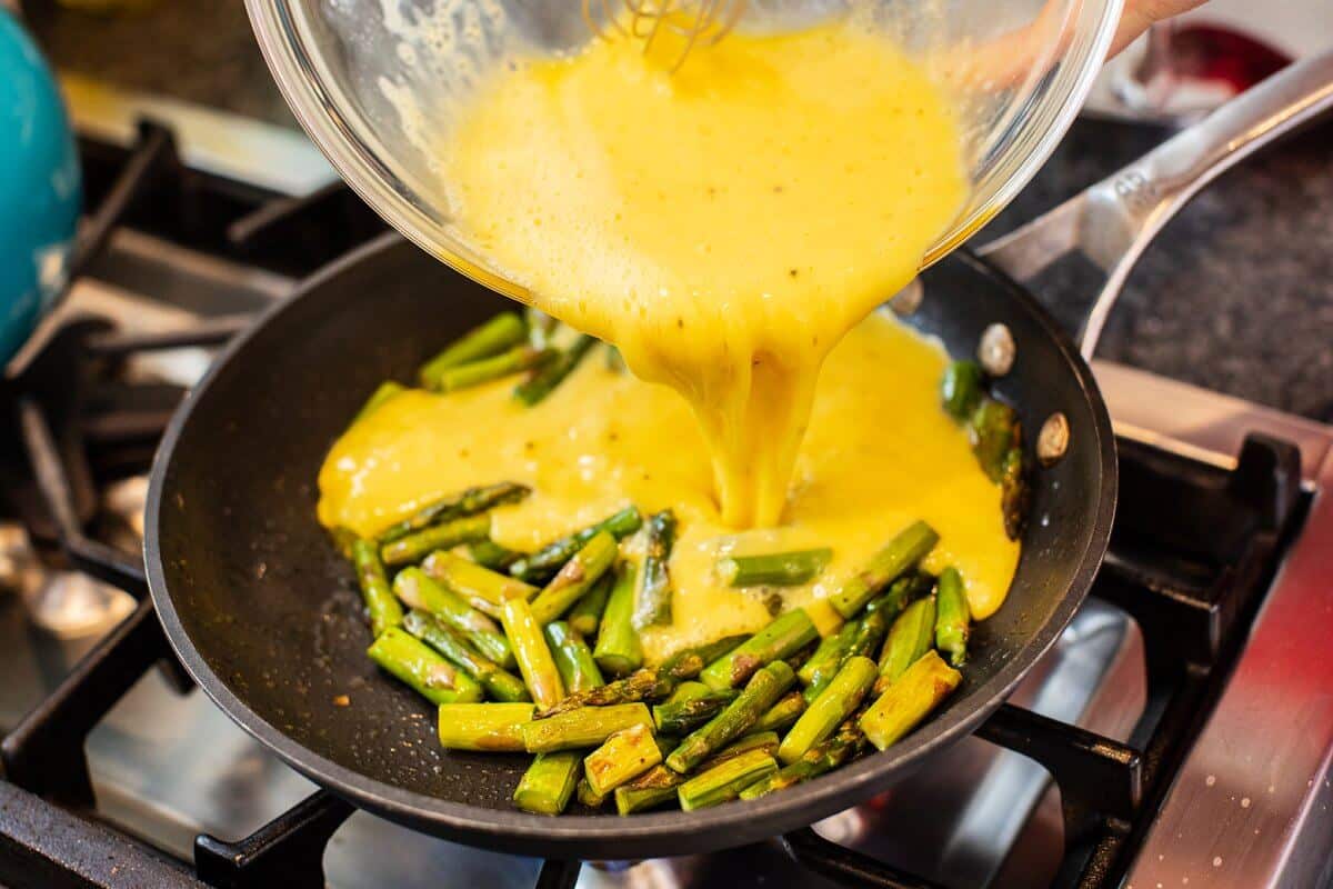 Pouring whisked eggs over the top of asparagus in skillet.