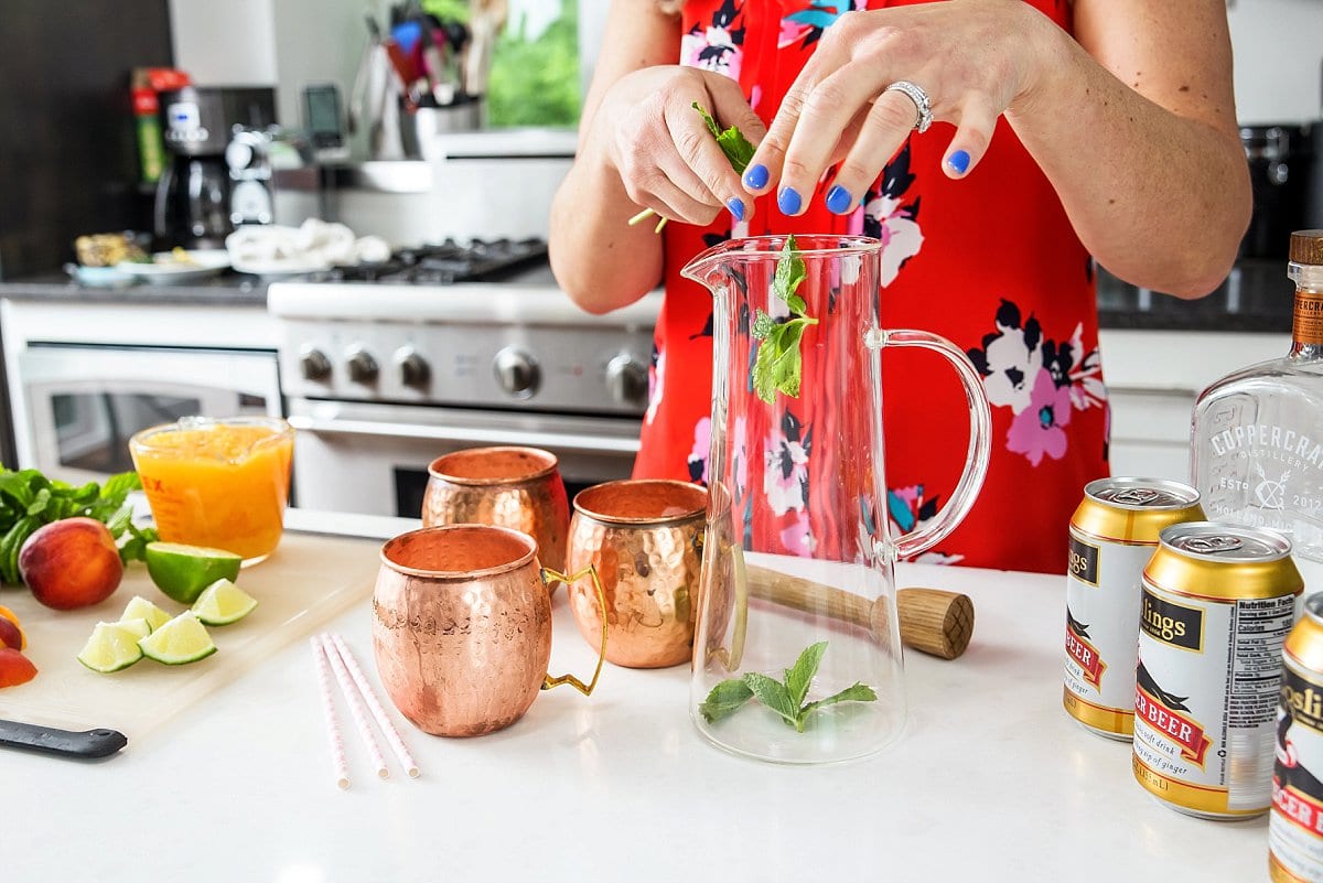 Adding mint leaves to a large pitcher on a counter next to copper mugs.