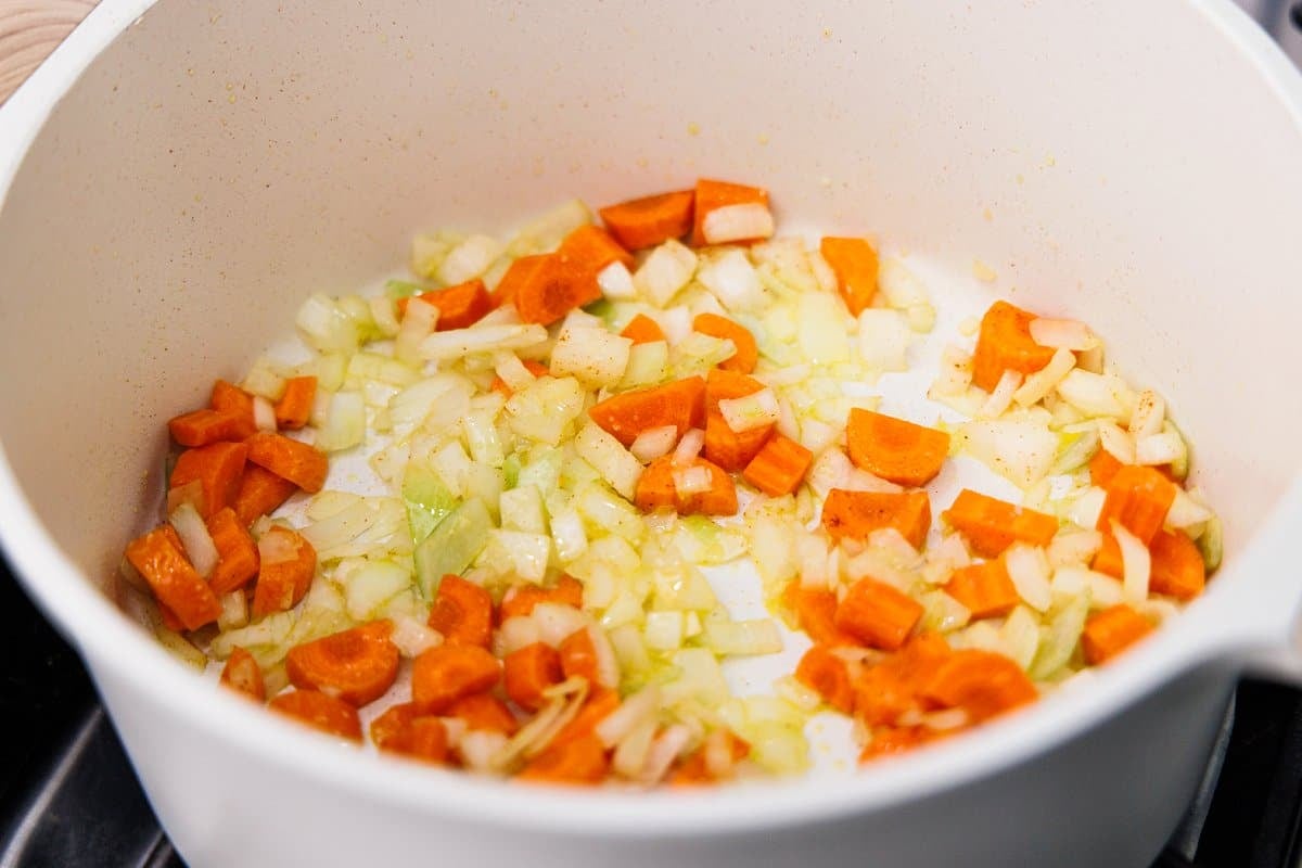 Sauteing carrots and onions in the bottom of a large white pot.