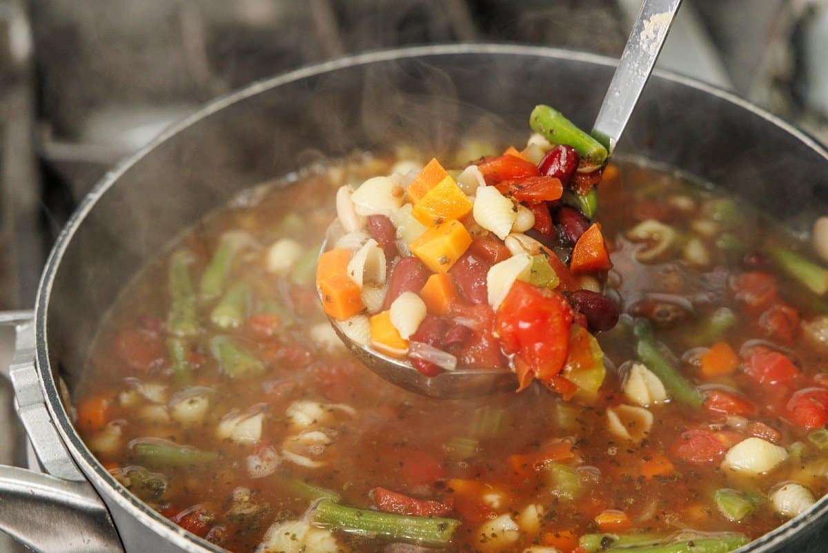 A ladle full of minestrone soup over a large soup pot.