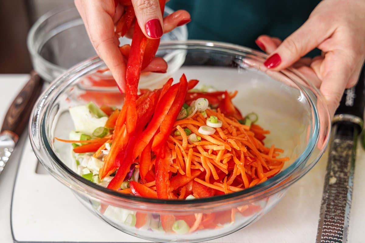 Putting sliced bell pepper in a bowl with shredded carrots and cabbage.