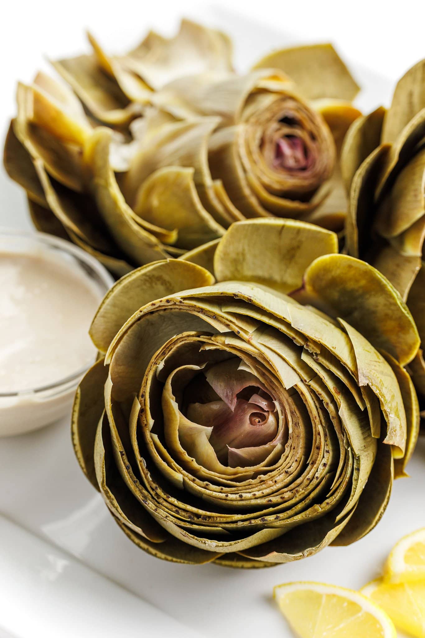 Steamed artichoke front and center, with more artichokes behind it and a small bowl of tahini dipping sauce.