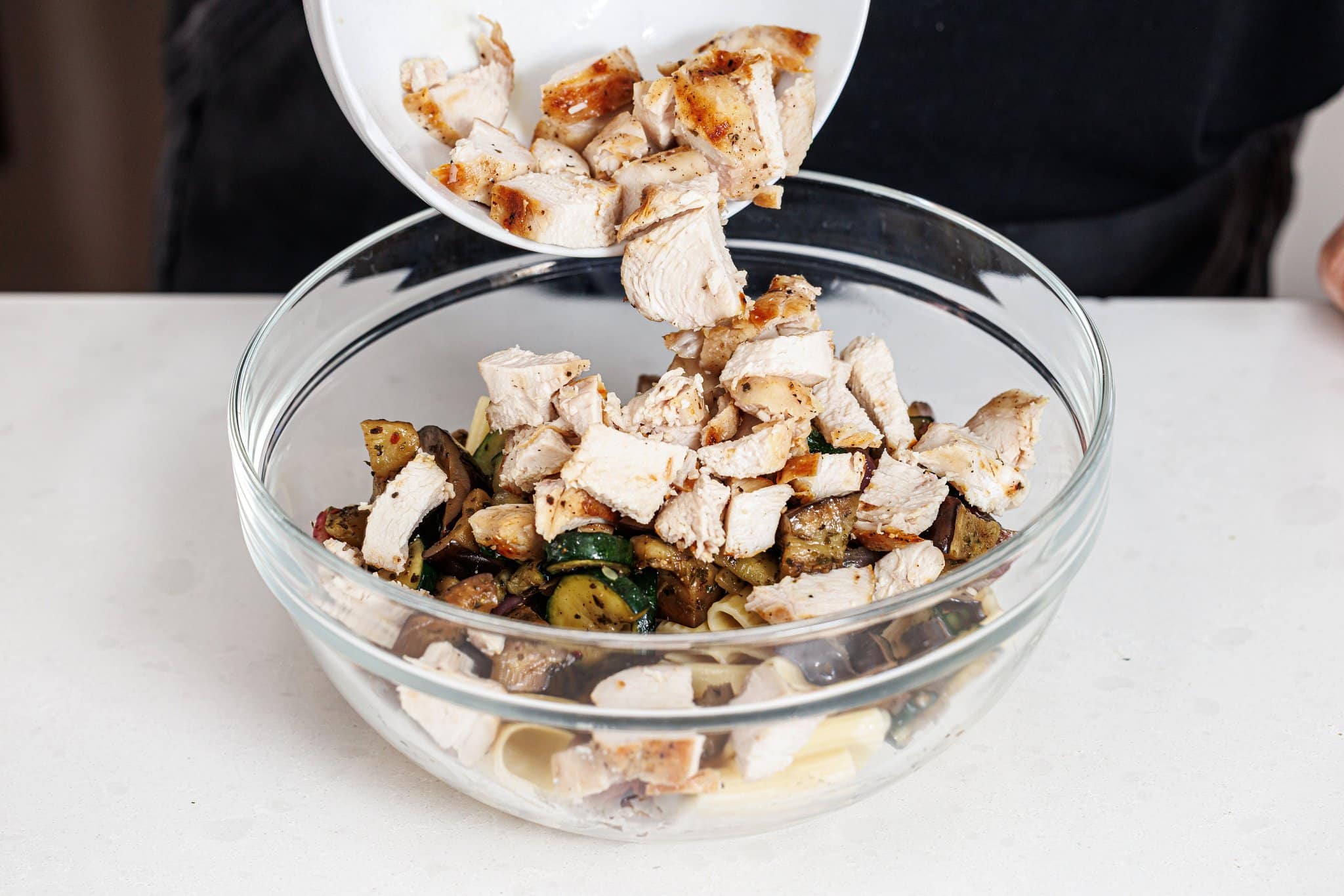 Adding cut up grilled chicken to large bowl of cooked pasta and veggies.