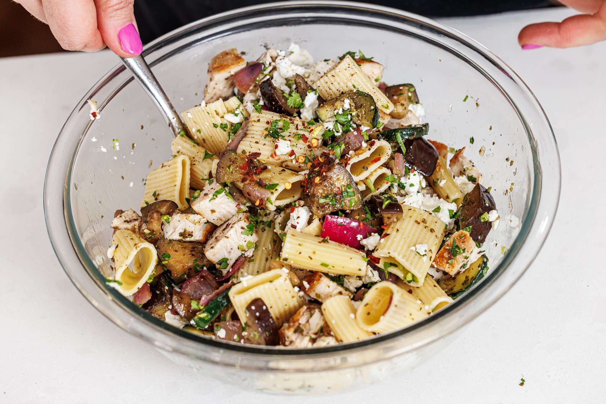 Tossing grilled chicken pasta salad together with a spoon in a glass bowl.