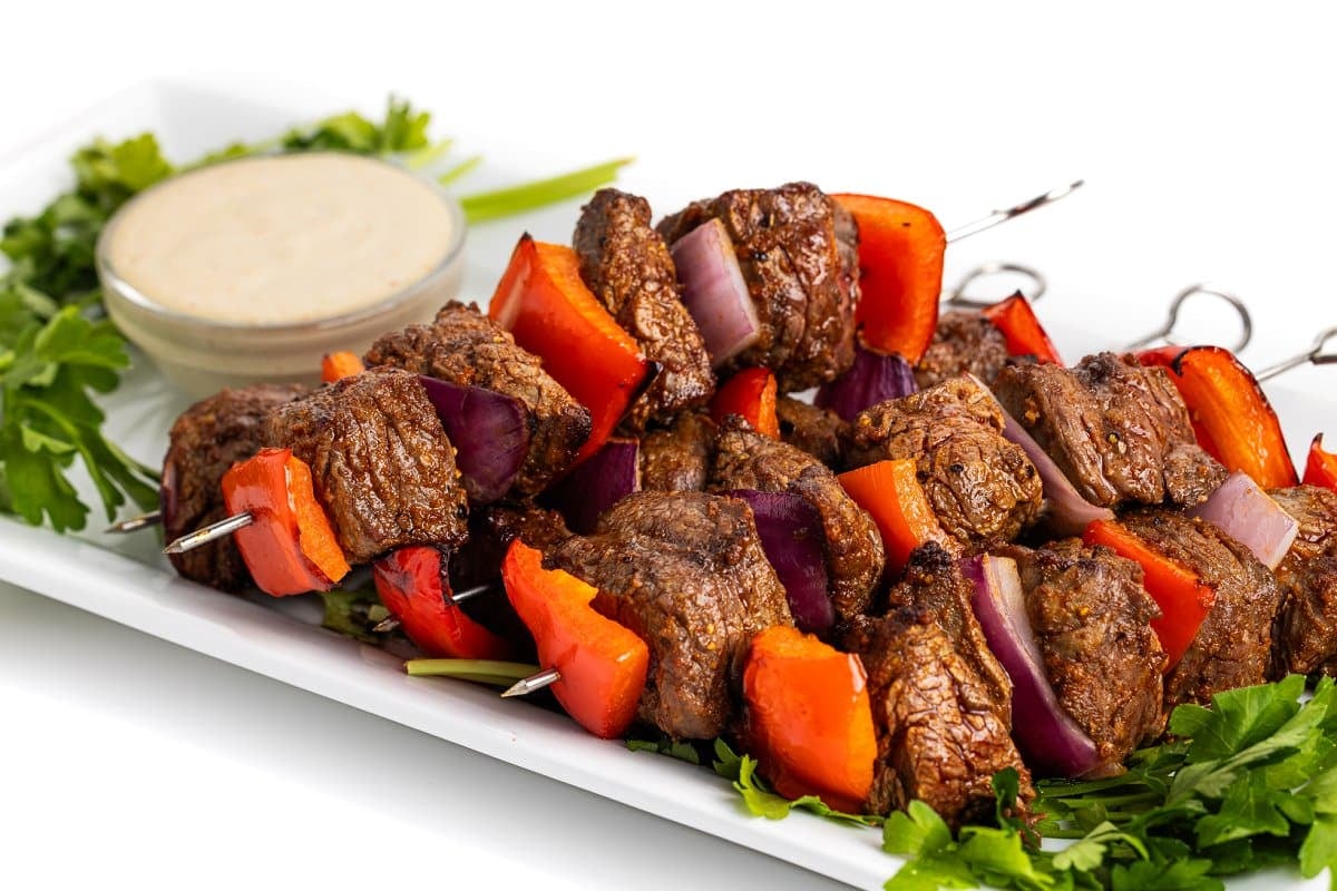 Steak skewers with tahini sauce on a platter with fresh parsley and broiled onion and peppers.