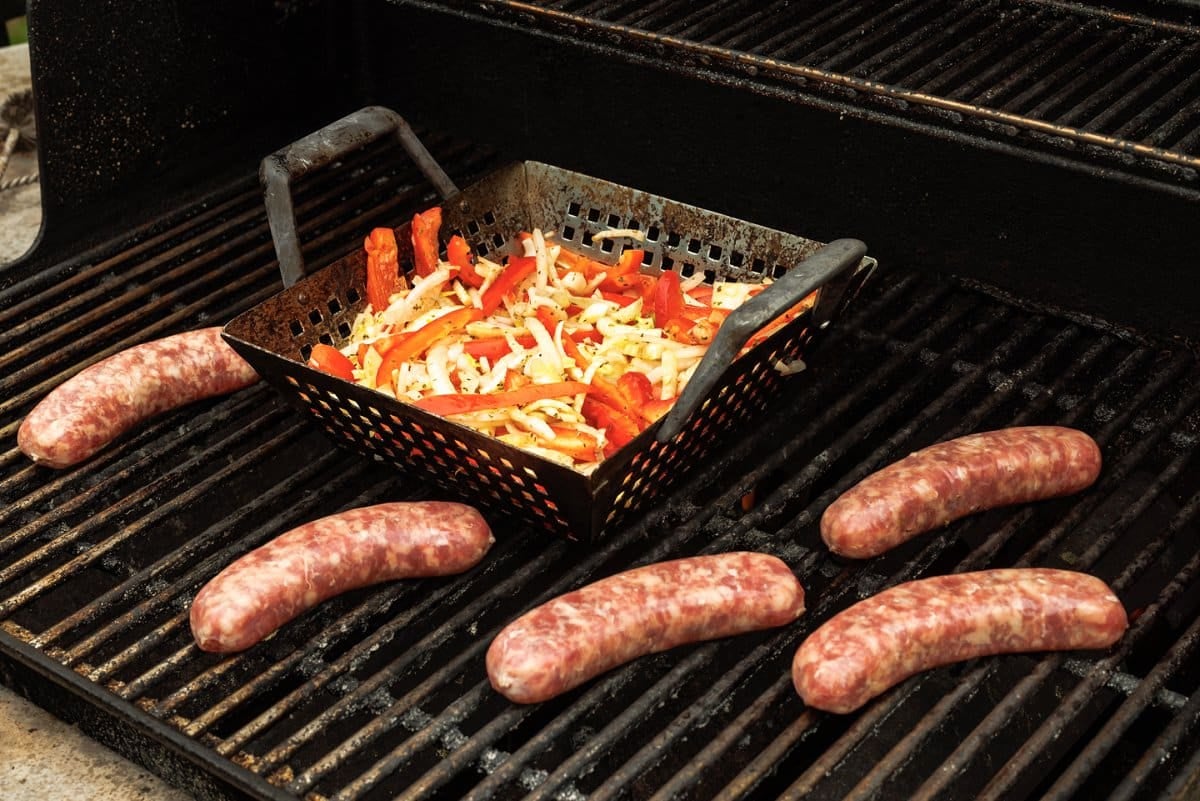 5 bratwursts on a grill with sliced peppers and onions in a grill basked in the center.