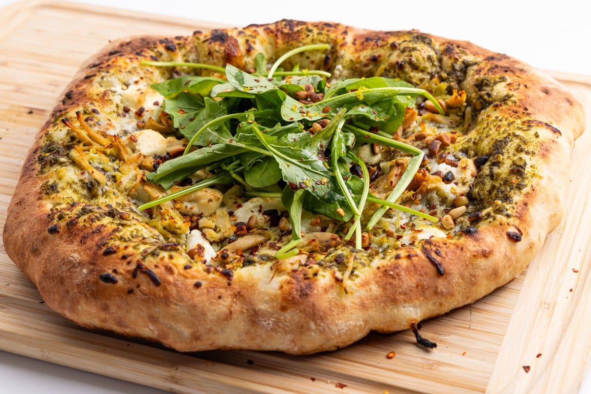 Cooked pesto chicken pizza with goat cheese and pine nuts on a cutting board.