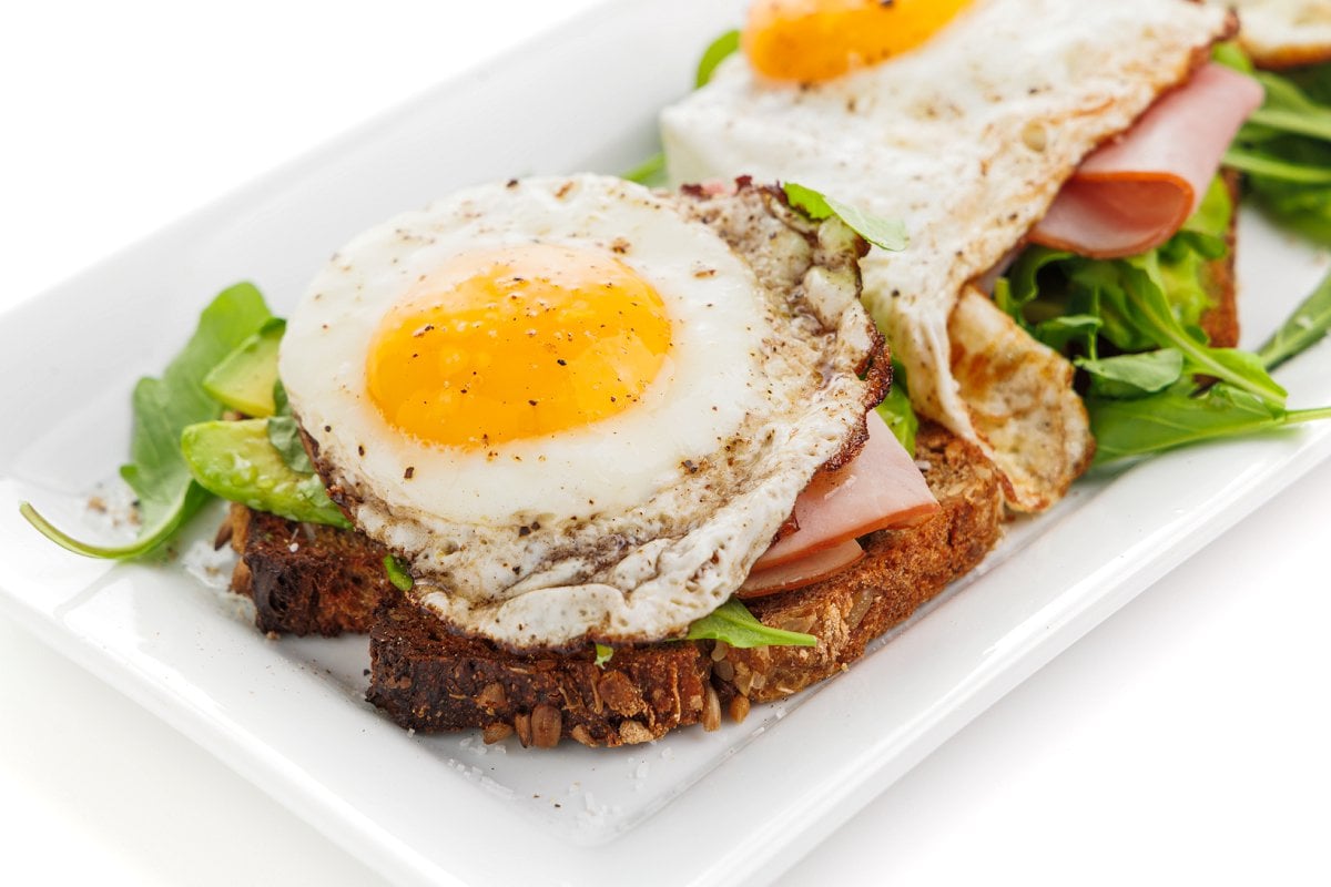 Avocado toast with egg, ham, and arugula seasoned with salt and pepper on a white plate.
