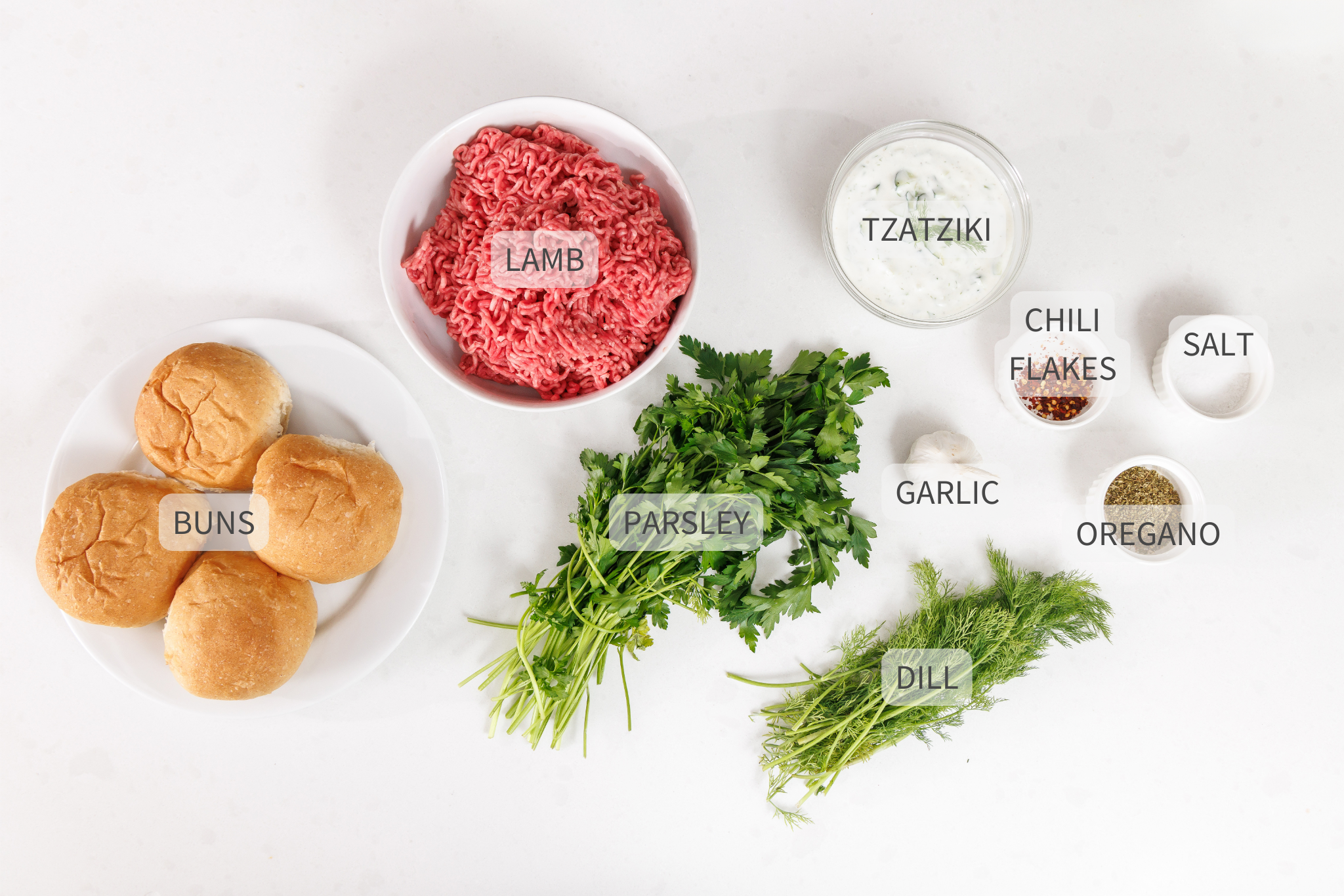 Labeled ingredients for greek lamb burgers with tzatziki sauce against a white counter.