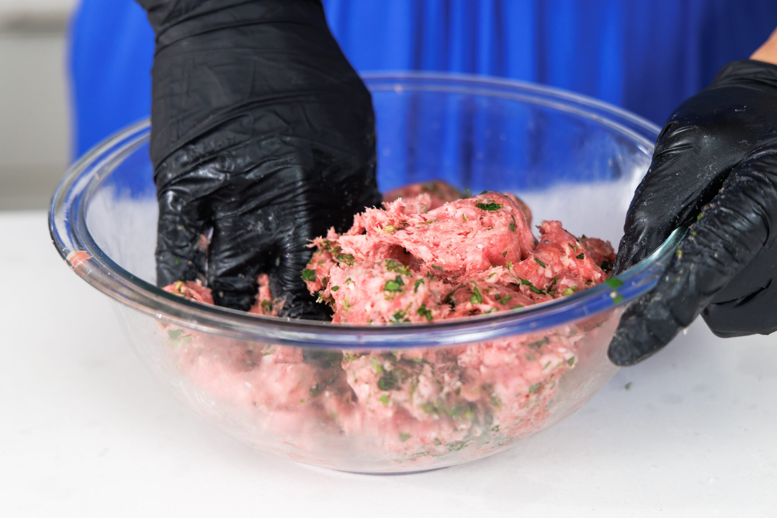 Hand mixing herbs into ground lamb in a glass bowl.