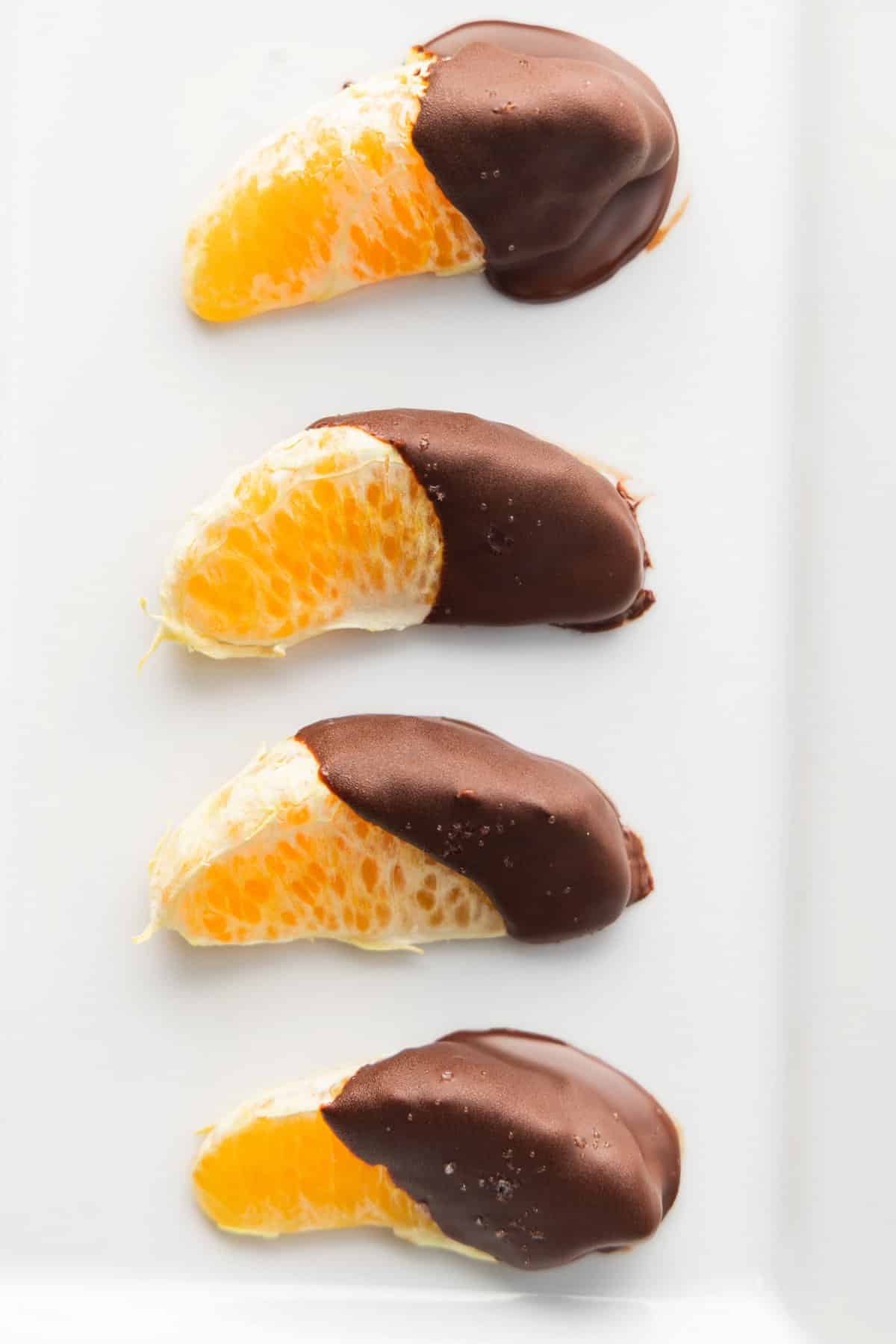 4 chocolate dipped clementines with sea salt in a row on a white rectangle plate.