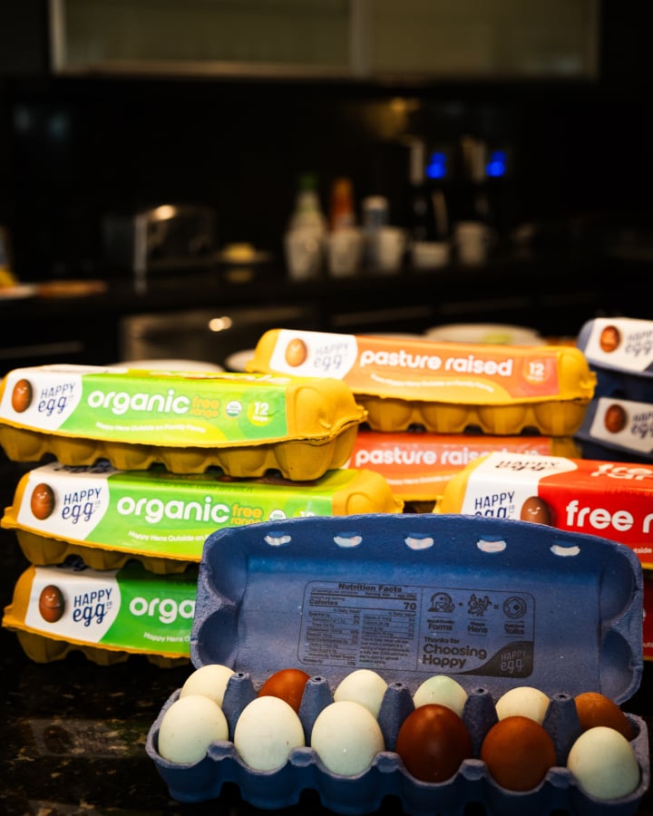 An open carton of Happy Egg eggs in front of six closed cartons of eggs.